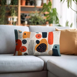 May include: Rectangular decorative pillow with an abstract pattern. The design features orange, navy, and yellow shapes with black accents on a cream background. The pillow is placed on a gray sofa, suggesting a home decor theme.