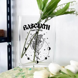 May include: Clear glass vase with a curved rectangular shape. The vase features a black graphic of a dragon wrapped around a sword, with the text "BASGIATH WAR COLLEGE". The vase holds green stems and white flowers.