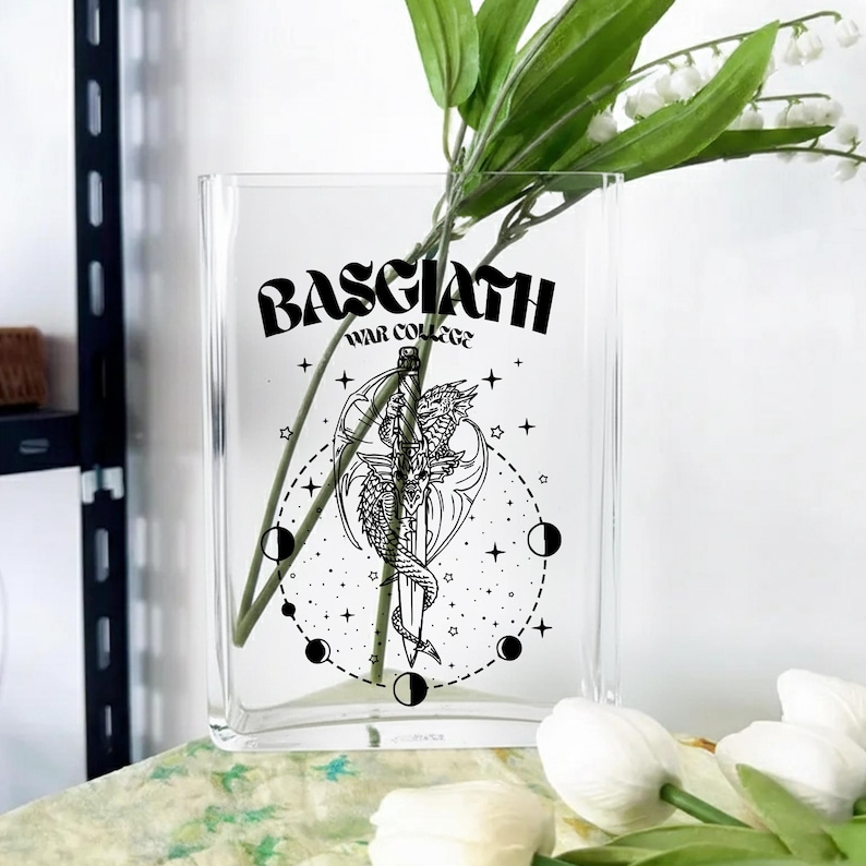May include: Clear glass vase with a curved rectangular shape. The vase features a black graphic of a dragon wrapped around a sword, with the text "BASGIATH WAR COLLEGE". The vase holds green stems and white flowers.
