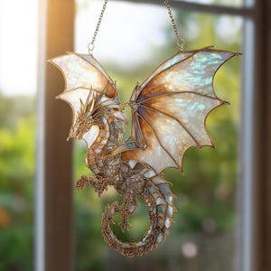 Puede incluir: Un atrapasoles de dragón de vidrieras con alas extendidas. El dragón presenta un diseño detallado con un marco dorado y alas iridiscentes en tonos blancos, marrones y azules. Cuelga de una delicada cadena.