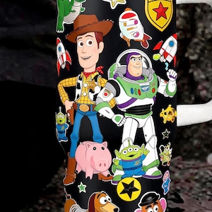 May include: A black stainless steel tumbler featuring a wrap-around graphic print of various animated characters including a cowboy, a space ranger, a green dinosaur, a pink pig, a dachshund, and a potato figure, with a white lid and handle.