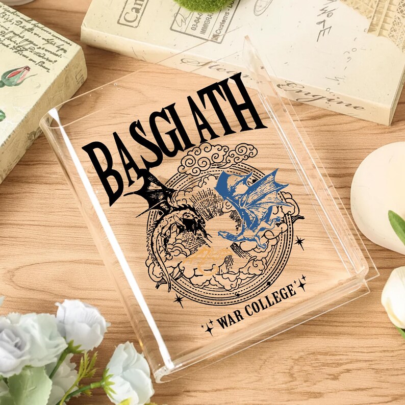 May include: A clear, rectangular book cover with the word "BASGIATH" in bold black letters. The cover features a circular design with two dragons, one black and one blue, and the text "WAR COLLEGE".