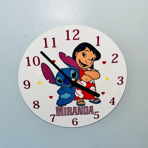 Può includere: Orologio rotondo bianco con i personaggi Lilo e Stitch. L'orologio ha lancette nere, numeri bordeaux e il nome "Miranda" stampato in basso. Sono inclusi anche cuori e stelle decorative.