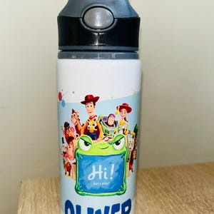Könnte beinhalten: Weiße Trinkflasche mit einem schwarzen und grauen Deckel, mit einem farbenfrohen Toy Story-Design. Die Flasche trägt den Namen "Oliver" in Blau, zusammen mit dem Text "Hi! Let's play!" und Bildern der Charaktere.
