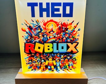 May include: A rectangular LED night light with the name "THEO" in blue block letters above the word "ROBLOX" in colorful letters. The light features a vibrant illustration of Roblox characters and a wooden base.