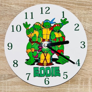 Könnte beinhalten: Eine weiße runde Uhr mit den Teenage Mutant Ninja Turtles. Das Zifferblatt hat grüne Zahlen und die Zeiger sind schwarz. Der Name "EDDIE" ist grün darunter gedruckt.