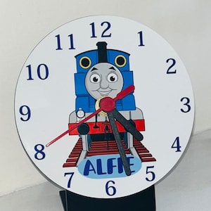 Könnte beinhalten: Eine runde Wanduhr mit einem blauen Zeichentrick-Lokomotiv-Charakter. Das Zifferblatt ist weiß mit schwarzen Zahlen und Zeigern. Der Name "ALFIE" ist auf einem blauen Banner unten aufgedruckt.