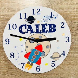Könnte beinhalten: Eine weiße, runde Wanduhr mit Weltraum-Motiv. Das Zifferblatt zeigt eine rot-blaue Rakete, Planeten, Sterne und den Namen "CALEB" in Blau. Schwarze Zeiger zeigen die Uhrzeit an. Die Zahlen sind blau.