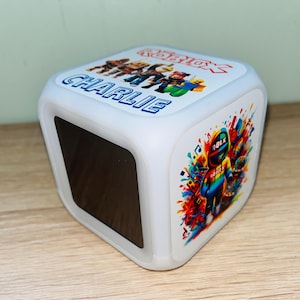 Może przedstawiać: Biała lampka nocna w kształcie kostki z postaciami z Roblox i imieniem "CHARLIE" w kolorze niebieskim. Jeden bok przedstawia kolorowy projekt postaci z Roblox z nutami. Na górze znajduje się logo Roblox i więcej postaci.