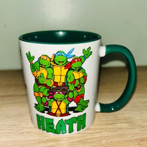 May include: White ceramic mug with a green interior and handle. The mug features a colorful graphic of the Teenage Mutant Ninja Turtles. The name "HEATH" is printed in green block letters at the bottom of the mug.