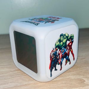 Op de afbeelding: Een witte, kubusvormige digitale wekker met Marvel-superhelden. De klok toont afbeeldingen van Hulk, Iron Man, Captain America en Thor. De bovenkant van de klok heeft de tekst "SUPER HEROES". De klok staat op een houten oppervlak.