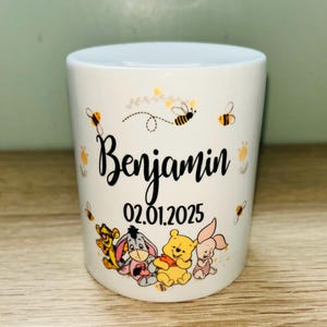 Könnte beinhalten: Weißer Keramikbecher mit dem Namen "Benjamin" in schwarzer Schrift und dem Datum "02.01.2025". Der Becher ist mit Illustrationen von Winnie-the-Pooh-Figuren und Bienen in Gelb und Schwarz verziert.