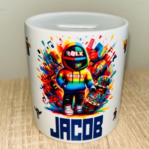 Könnte beinhalten: Weiße Keramik-Spardose mit einem farbenfrohen Roblox-Charakterdesign und dem Namen "JACOB" in blauen Blockbuchstaben. Das Design umfasst einen Regenbogen-Hoodie, ein Skateboard und einen lebendigen Farbspritzer.
