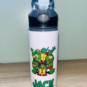 Può includere: Borraccia bianca con coperchio nero e grigio e beccuccio ribaltabile. La borraccia presenta una grafica dei Teenage Mutant Ninja Turtles e il nome "JACK" in lettere verdi.