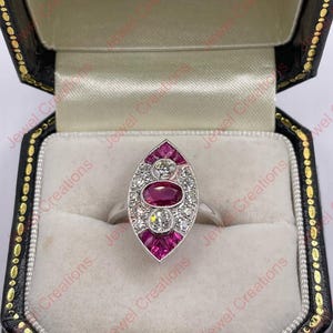 May include: A silver ring with an oval design, set with a central ruby and surrounded by smaller rubies and clear gemstones. The ring is displayed in a black jewelry box with a gold clasp and a white interior.