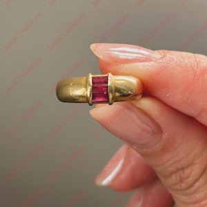 Puede incluir: Un anillo de oro con tres piedras preciosas rectangulares de color rojo intenso en el centro. El anillo tiene una banda redondeada y un acabado pulido. El anillo se sostiene entre los dedos sobre un fondo neutro.