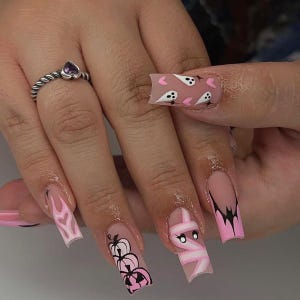 May include: A silver ring with a purple heart-shaped stone is on a finger next to long, square acrylic nails. The nails feature Halloween designs, including ghosts, pumpkins, a mummy, and a bat, in pink, black, and white.