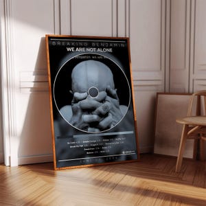 May include: A rectangular wall art print featuring the Breaking Benjamin album We Are Not Alone. The black and white design displays a person with hands covering their face, a tracklist, and the release date of June 29, 2004, in a wooden frame.