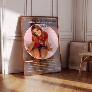 Peut inclure: A framed poster of Britney Spears' album "...Baby One More Time." The poster is in a brown wooden frame with a grey background. The album cover art features Britney in a red top and denim skirt. The tracklist and release date are also visible.