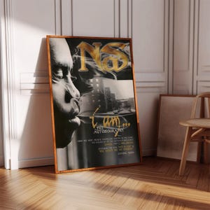 May include: A framed poster with a wooden frame, featuring a black and white profile of a person. The poster has gold text reading "Nas" and "I Am... The Autobiography". It is leaning against a wall.
