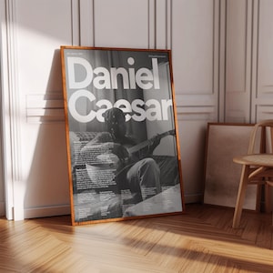 May include: A rectangular poster in a wooden frame featuring a black and white photograph of a person playing an acoustic guitar. The text "Daniel Caesar" is printed in large white sans-serif letters at the top, with smaller biographical text below.