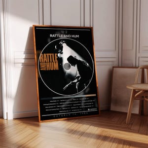 Może przedstawiać: Oprawiony plakat z albumem U2 "Rattle and Hum". Plakat ma czarne tło z okrągłym zdjęciem gitarzysty. Tytuł albumu jest w kolorze pomarańczowym. Plakat zawiera listę utworów i datę 10 października 1988 r. Rama jest drewniana.