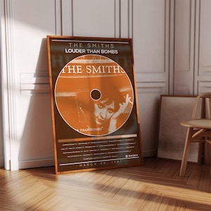Pode incluir: Um poster emoldurado com a arte do álbum "Louder Than Bombs" dos Smiths. O design inclui uma imagem circular em tons de laranja, com o nome da banda e o título do álbum em branco. O poster é datado de 30 de março de 1987.