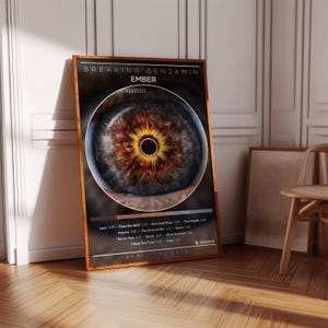 May include: A rectangular poster in a wooden frame featuring the Breaking Benjamin album Ember. The design displays a large, detailed human eye with a brown and gold iris, a tracklist, and the release date April 13, 2018, on a dark background.