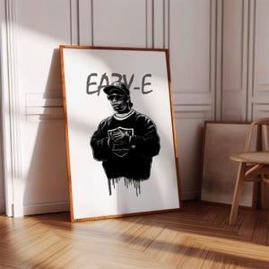 Puede incluir: Retrato en blanco y negro enmarcado de Eazy-E, con gorra y gafas de sol negras. La obra presenta el texto "EAZY-E" sobre la figura y un diseño de escudo en la sudadera. El marco es de color marrón cálido.