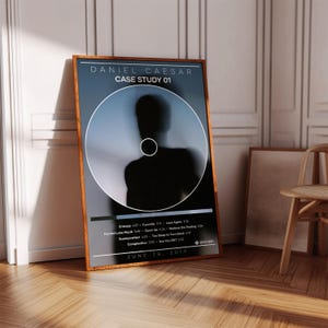 May include: A rectangular wall art print in a wooden frame featuring the Daniel Caesar Case Study 01 album cover. The design includes a central circular graphic with a dark silhouette, track list text, and the release date June 28, 2019.