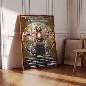 Puede incluir: Un póster enmarcado con el álbum "Oops!... I Did It Again" de Britney Spears. El póster tiene un marco de madera marrón y muestra la portada del álbum, la lista de canciones y la fecha "MAY 3, 2000". La portada del álbum presenta una foto de Britney Spears.