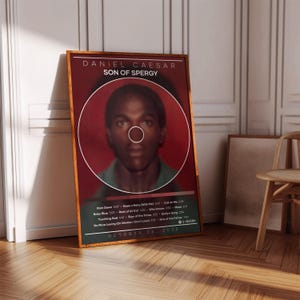 May include: A framed poster featuring a portrait of Daniel Caesar, with the text "SON OF SPERGY" above the image. The poster has a dark red background with a white circular outline around the portrait. Below the image are song titles and the date "OCTOBER 24, 2025".