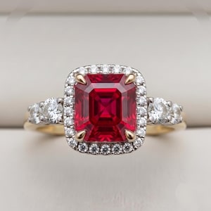 Può includere: Un anello con una grande pietra preziosa rossa rettangolare taglio smeraldo incastonata in oro giallo. La pietra centrale è circondata da un alone di piccoli diamanti bianchi rotondi. Due diamanti bianchi rotondi aggiuntivi su ogni lato.