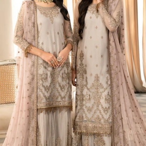 May include: Two women in matching light grey and gold embroidered outfits. Each outfit features a long-sleeved tunic, matching trousers, and a sheer, pale pink scarf. The outfits are adorned with detailed gold embroidery and delicate fringe.
