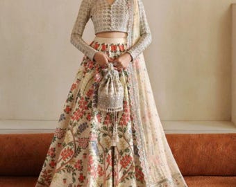 Pode incluir: Um lehenga marfim com padrão floral, blusa de manga comprida e dupatta. A saia e a blusa apresentam bordados e enfeites detalhados. Uma pequena bolsa de contas é segurada. Medidas em centímetros.