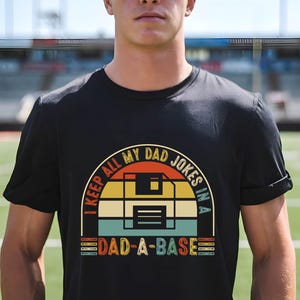Op de afbeelding: Zwart T-shirt met een retro design. Het ontwerp heeft een kleurrijke boog met de tekst "I KEEP ALL MY DAD JOKES IN A DAD-A-BASE". Het ontwerp bevat een floppy disk-afbeelding. Het shirt wordt door een persoon gedragen.