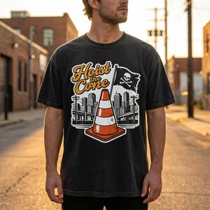 Pu&ograve; includere: T-shirt nera con una grafica raffigurante un cono stradale, un paesaggio urbano e una bandiera pirata. Il testo "Hoist the Cone" &egrave; in un font stilizzato. Il design &egrave; in bianco, arancione e nero.