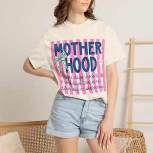 Puede incluir: Camiseta color crema con la palabra "MOTHERHOOD" en letras azules sobre un fondo de rayas rosas. La camiseta también presenta el texto "some days I rock it, some days it rocks me, either way we are rocking."