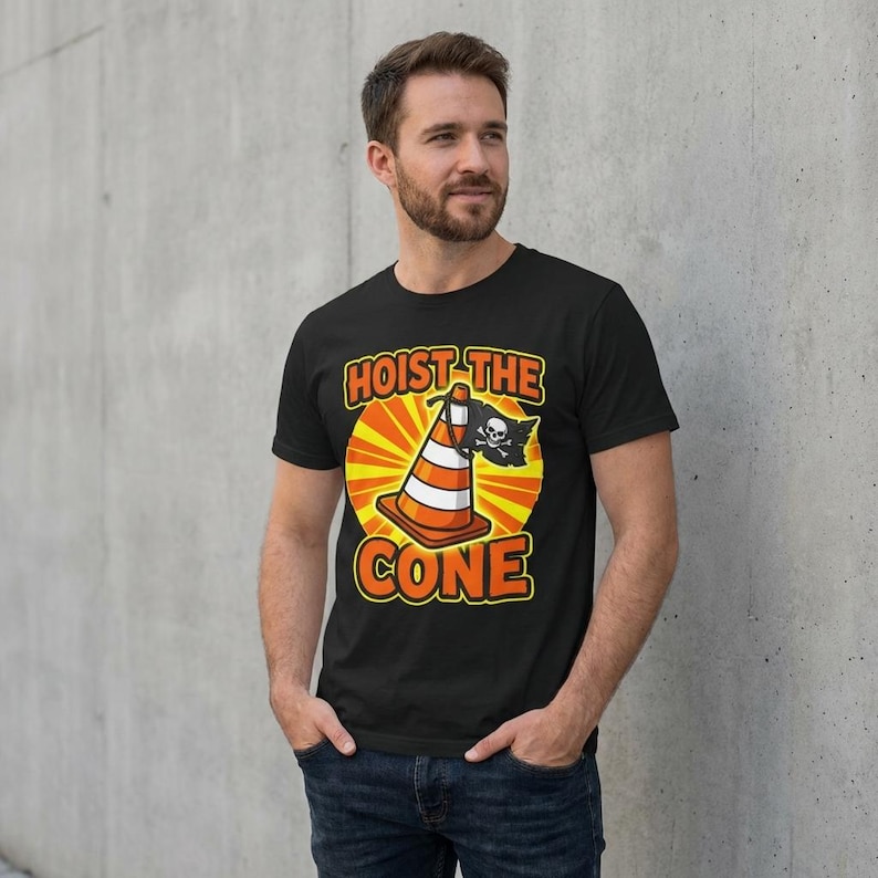 Puede incluir: Camiseta negra con un dise&ntilde;o de cono de tr&aacute;fico y elementos tem&aacute;ticos piratas. El texto "HOIST THE CONE" est&aacute; en naranja y blanco. El dise&ntilde;o incluye una calavera y huesos cruzados.