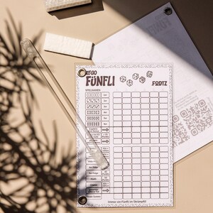 Könnte beinhalten: Ein transparentes Acryl-Spielergebnisblatt mit dem Titel FÜNFLI mit einem Raster für Würfelwürfe, begleitet von einem langen, transparenten Acryl-Stift und zwei kleinen, rechteckigen weißen Radiergummis. Das Blatt enthält Würfelsymbole und einen QR-Code.