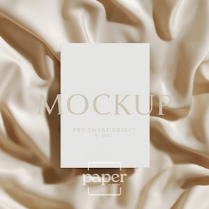 May include: A white rectangular mockup card with the word "MOCKUP" in a light brown font. The card is on a beige satin fabric background. The text "PSD SMART OBJECT + JPG" is below the word "MOCKUP". The logo "paper in mind" is at the bottom.