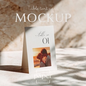 May include: A white table tent card mockup with the text "MOCKUP" and "Table tent card" in a light font. The card features a photo of a couple kissing at sunset and the text "table no 01". The logo "paper in mind" is at the bottom.