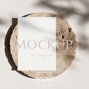 May include: A white rectangular mockup card with the word "MOCKUP" printed on it, resting on a textured, circular, beige stone. The card also has the text "PSD SMART OBJECT + JPG" printed on it. The word "paper" is printed at the bottom.
