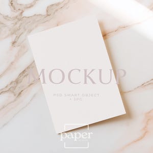 May include: A blank, off-white rectangular card with the word "MOCKUP" in light purple text. Below the word "MOCKUP" is the text "PSD SMART OBJECT + JPG". The card is on a marble surface. The logo "paper" is in the bottom right corner.