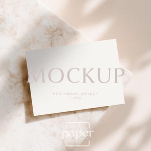 Op de afbeelding: Een witte rechthoekige kaart met het woord "MOCKUP" in lichtroze letters. Daaronder staan de woorden "PSD SMART OBJECT + JPG". De kaart ligt op een marmeren oppervlak met een schaduw.