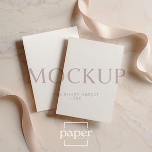 May include: Two blank white rectangular cards arranged in an overlapping vertical orientation. The top card features the text MOCKUP, PSD SMART OBJECT + JPG. A light beige ribbon curves across the composition, which has a marble-like texture.