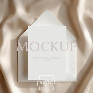 May include: A white mockup card and envelope set against a beige satin background. The word "MOCKUP" is printed in a light purple color across the card. The text "PSD SMART OBJECT + JPG" is printed below the word "MOCKUP". The word "paper" is printed at the bottom.