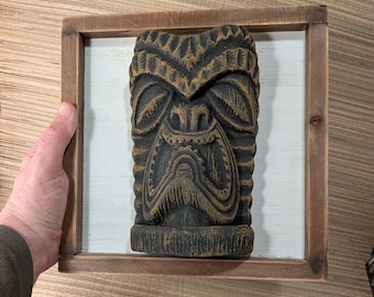 May include: A carved wooden tiki mask in a brown wooden frame. The mask is dark brown with gold accents, and is mounted on a white background. The frame is a light brown color.