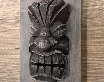 May include: A dark brown wooden wall plaque featuring a carved tiki mask with a distressed finish. The mask has a prominent forehead, deep-set eyes, a wide nose, and a mouth with visible teeth. The plaque has a scalloped top and bottom edge.