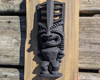 May include: A carved wooden tiki statue in black, mounted on a rectangular wooden plaque with a scalloped top edge. The plaque has a light brown finish with orange accents. The tiki has a detailed face and body.
